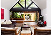 View from interior with seating area through triangular, glazed gable to terrace with pergola and tropical garden