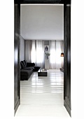 View through black double door into purist living room with glossy, white floorboards and dark sofa in front of curtains at windows