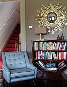 Blue armchair next to half-height bookcase below mirror with sunburst gilt frame on wall