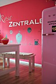 Deep pink wall with white lettering and pale pink retro fridge in foreground