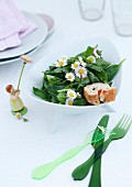 Green plastic cutlery and ornament of girl next to bowl of spinach salad with daisies