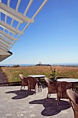 Rattanstühle und Tische auf sonnenbeschienener Terrasse unter Lamellen-Vordach vor weitläufiger Landschaft