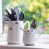 Large and small items of cutlery in two old china jugs with floral patterns in front of window