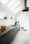 Monolithic kitchen counter with black fronts below strip of skylights and metal, industrial-style pendant lamps