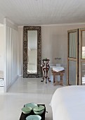 Vintage bowls on trunk at foot of bed and floor-to-ceiling framed mirror leaning against wall next to bathtub in bathroom area