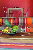 Sitzplatz mit bunt gestreiften Polstern und Schale mit tropischen Früchten auf Holztisch; Esstisch vor rot getönter Wand im Hintergrund