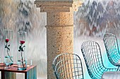 Stühle aus Drahtgestell und Rosen in Glasvasen auf Beistelltische vor Steinsäule und wasserfallartiger Wand