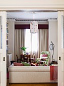 View through open sliding doors of pale couch and floral armchair in lounge area in front of windows with floor-length curtains