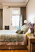 Double bed with elegant bedspread and matching scatter cushions next to open balcony door in traditional, elegant bedroom