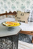 Dish of sliced citrus fruits on table and wooden bench with carved backrest against floral, fabric wallpaper
