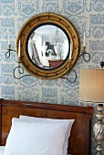 Runder gerahmter Spiegel mit Kerzenhaltern an tapezierter Wand über dem Bett mit Kopfteil aus Holz