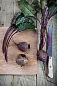 Beetroot and beetroot leaves on a chopping board, and a knife on a wooden surface