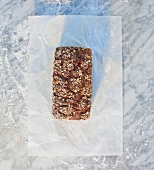 A tin-baked loaf topped with rolled oats, pumpkin seeds and sunflower seeds, on paper, on a marble slab dusted with flour