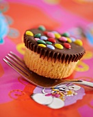 A cupcake decorated with chocolate glaze and colourful chocolate beans