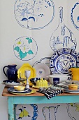 Geschirr in Blau, Weiß und Gelb auf einem Tisch vor Wandposter mit Geschirrmotiv in den gleichen Farben
