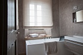 Designerbad - Blick durch offene Tür auf Badewanne vor Fenster mit heruntergezogenem Rollo und moderner Waschtischtrog an Wand