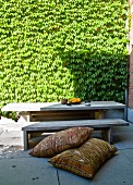 Folkloristische Kissen auf Boden vor rustikaler Holzbank und Tisch in sonnenbeschienenem Innenhof mit begrünter Wand