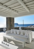 Cushions arranged on sofa with white loose cover and minimalist table and bench on roofed terrace in front of open sea