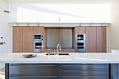 Central island with white worksurface and integrated sink in front of kitchen cupboards with wooden doors and fitted appliances