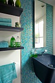 Waschbecken aus schwarzem Mineralwerkstoff und Spiegel an türkiser Mosaikfliesenwand neben Wandbords mit Badutensilien