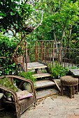Alter Rattanstuhl neben Holztreppe und rostigem metall Geländer in dicht bewachsenem Garten