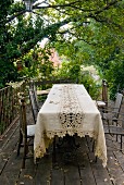 Tablecloth with lace insert and metal chairs on wooden terrace in garden with dense vegetation