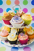 close up of purple, yellow and pink cup cakes decorated with icing flowers on a purple pansy flowered cake standon a coloured spotty background