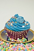 close up of single blue iced cup cake decorated with sprinkles and sweets with a white background