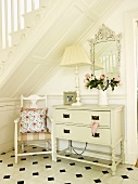 Chair with cushion, lamp and vase of flowers on white-painted chest of drawers and wall mirror below wooden staircase in hall