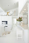 White, modern interior with bar stools at kitchen counter opposite lounge area with collection of swords and shields on top of fitted cupboards