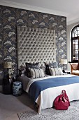 Prunkvolles Bett mit gestepptem, hohen Kopfende an taubenblauer Wandtapete mit floralem Muster; auf dem Bett Zierkissen Ton in Ton mit der Wandtapete