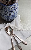 Cutlery holder with a blue and white design; to one side, a lace-edged napkin and three antique silver spoons