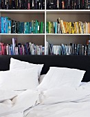 Bookcase organised by book colour against grey wall; untidy white bed linen on bed with black headboard in foreground