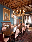 Opulent, long wooden table in front of marble fireplace in lavish dining room with elaborately carved coffered ceiling