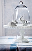 Two white china rabbits on cake stand with glass cover