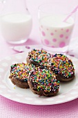 Chocolate pikelets with colourful sugar sprinkles, with glasses of milk in the background