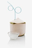 A peeled coconut with a drinking straw and a small bowl of coconut water