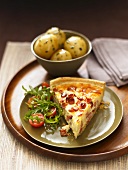 A slice of quiche lorraine with rocket salad and a side dish of potatoes