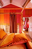 Pillows in yellow pillowcases on four-poster bed with wooden frame opposite wall with stripes of various shades of red in rustic bedroom