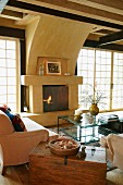 Alte Holztruhe zwischen Couch und Sessel um Glastisch und offener Kamin in Wohnzimmer mit Sprossen Terrassentüren