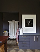 Dark sideboard against partition of same colour with modern artwork on white mount next to open doorway showing view of white farmhouse cupboard in room beyond