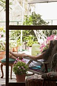 Blick durch Fenstertür auf begrünte Terrasse - Frau im Sonnenstuhl mit auf Antikhocker hochgelegten Beinen