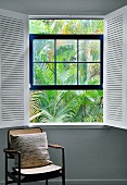 Pillows on an antique chair by a window with open, white interior shutters and a view of palm fronds