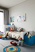 Kinderzimmer mit Spielsachen auf Teppichboden und blauem Sitzwürfel am Bettende vor grauer Wand
