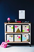 Storage boxes decorated with colourful triangles on retro-style shelving unit on castors against dark wall