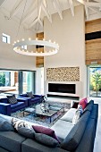 Large corner sofa and armchairs beneath a contemporary ring pendant light in a two story living room with hipped roof; ceiling height fireplace with firewood storage