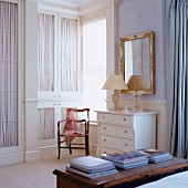 Stacked books on trunk, fitted wardrobes with curtains behind glass door panels and white chest of drawers below mirror on wall in traditional interior