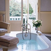 Badewasser in einer Vintage Badewanne mit weißem Rosenstrauss auf Ablage und Blick durch halboffene Terrassentür in den Garten