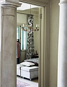 View between two pillars into bedroom with walls painted pastel green and chandelier; two ottomans on castors as bedroom bench