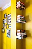 Pictures and clocks on yellow wall separated from floating bookshelves in niche by pillar